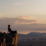 A hiker sitting on a rocky summit overlooking the Phoenix skyline at sunset, with desert mountains on the horizon.