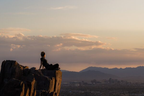 A hiker sitting on a rocky summit overlooking the Phoenix skyline at sunset, with desert mountains on the horizon.