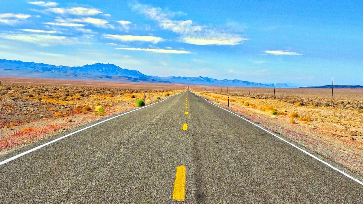 Open two-lane highway through the Nevada desert with mountains on the horizon under a bright blue sky.