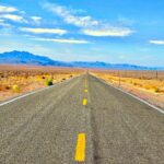 Open two-lane highway through the Nevada desert with mountains on the horizon under a bright blue sky.