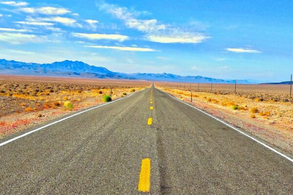 Open two-lane highway through the Nevada desert with mountains on the horizon under a bright blue sky.